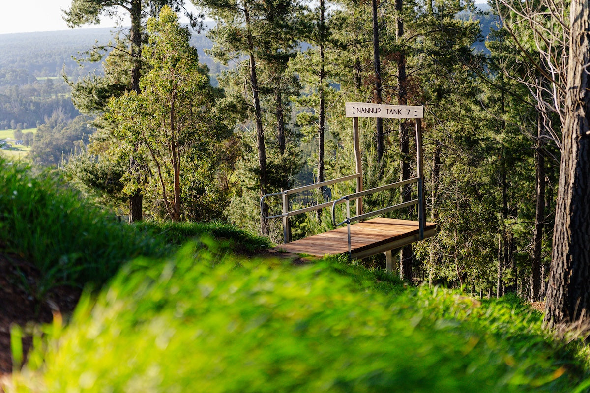 Green forest with a mountain bike jump and sign stating Nannup Tank 7.