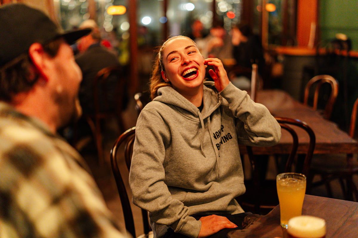 Two people sitting at a table, laughing and smiling in Nannup Brewery with a pint of beer, wearing a grey Nannup Brewing hooded jumper.