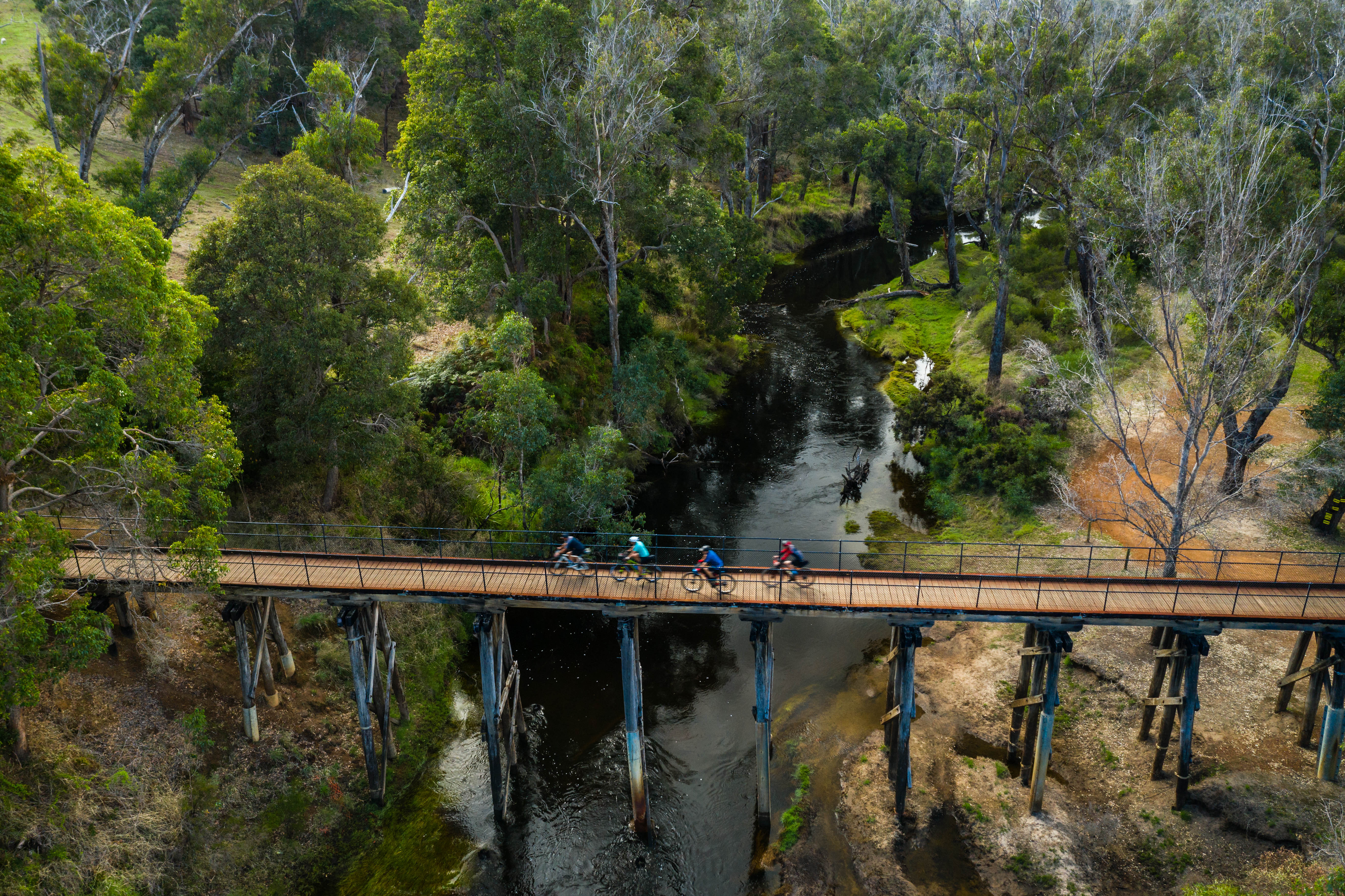 An aerial view of a wooden boardwalk bridge spanning a narrow river, with three cyclists riding across it. The surrounding area is dense green forest.