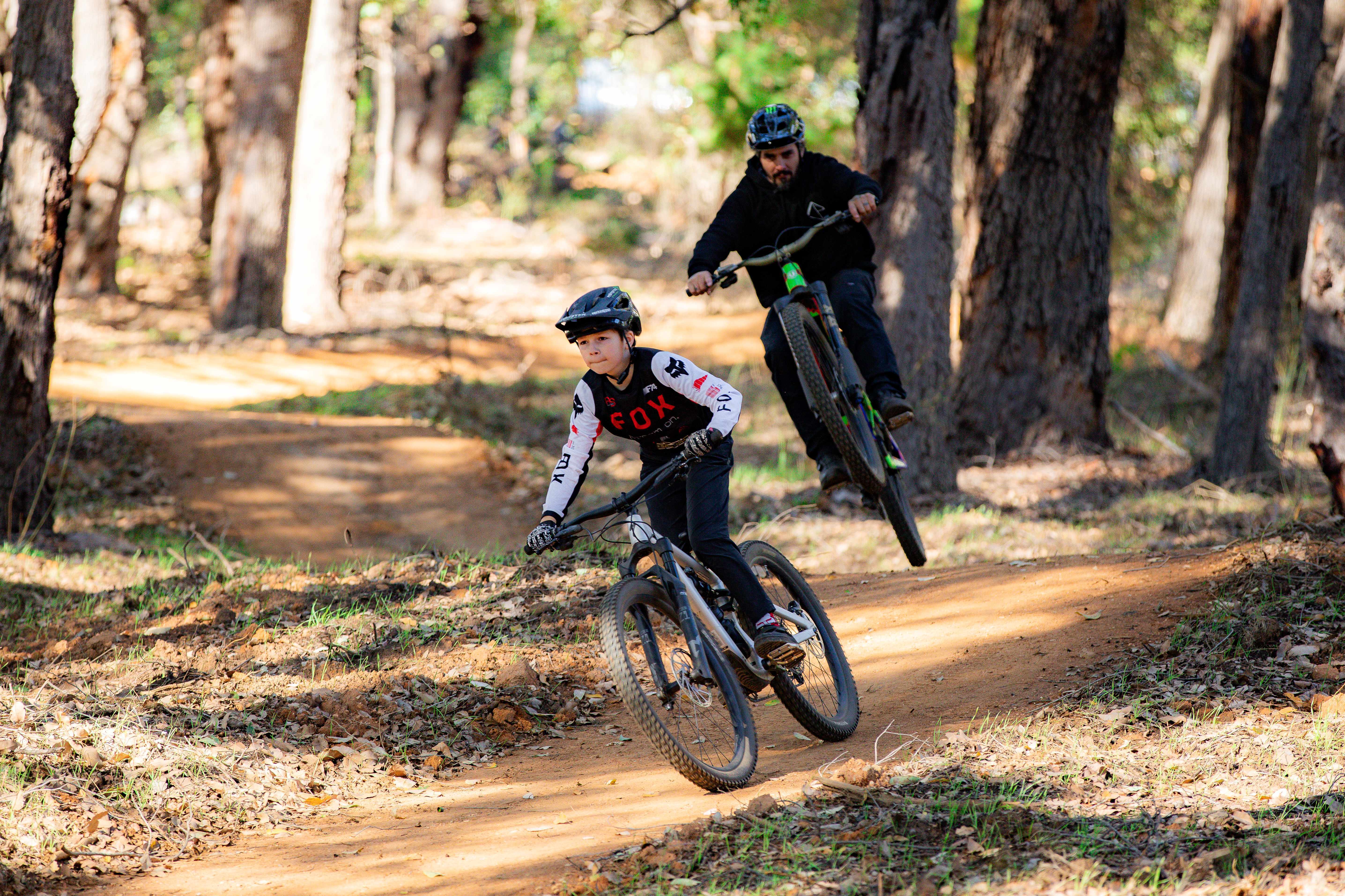 Two mountain bikers riding on a dirt trail through a wooded forest. The rider in the foreground wears a black helmet and a white, black, and red jersey, while the rider behind wears a black helmet and black clothing.